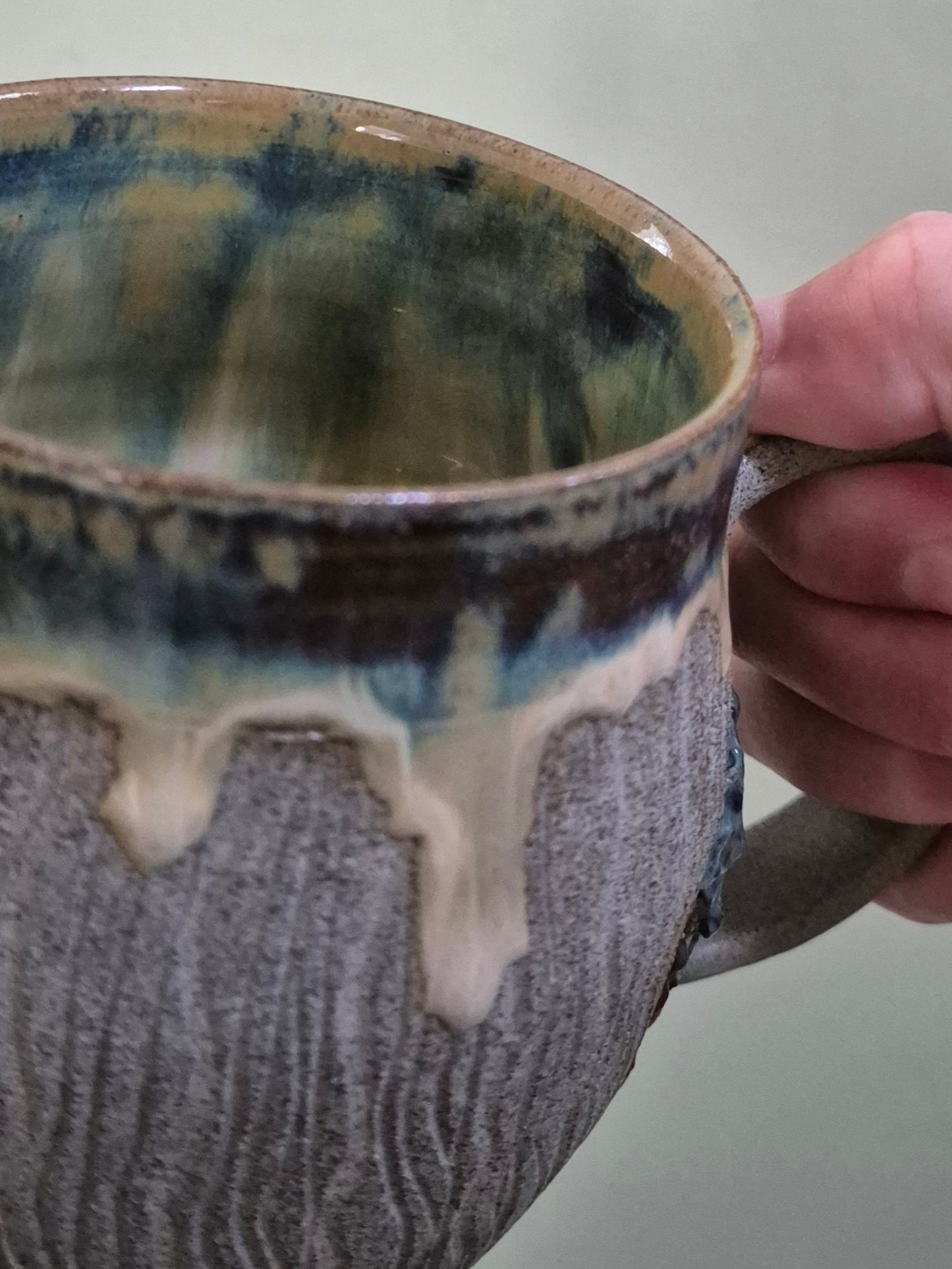 A close-up of a handmade ceramic mug with a textured gray exterior and a dripping glaze of blue, green, and cream on the inside, held by a hand against a neutral background.