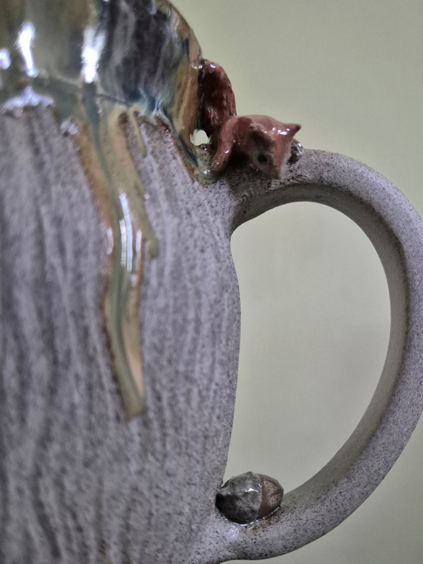 Close-up of a handmade ceramic mug with a textured gray-brown glaze, a small brown squirrel figurine perched on the rim, and a glossy amber drip of glaze near the top, set against a soft beige background.