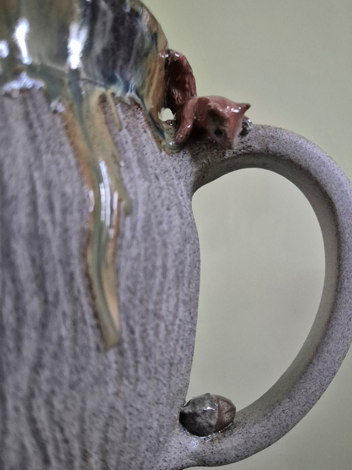 Close-up of a handmade ceramic mug with a textured gray-brown glaze, a small brown squirrel figurine perched on the rim, and a glossy amber drip of glaze near the top, set against a soft beige background.