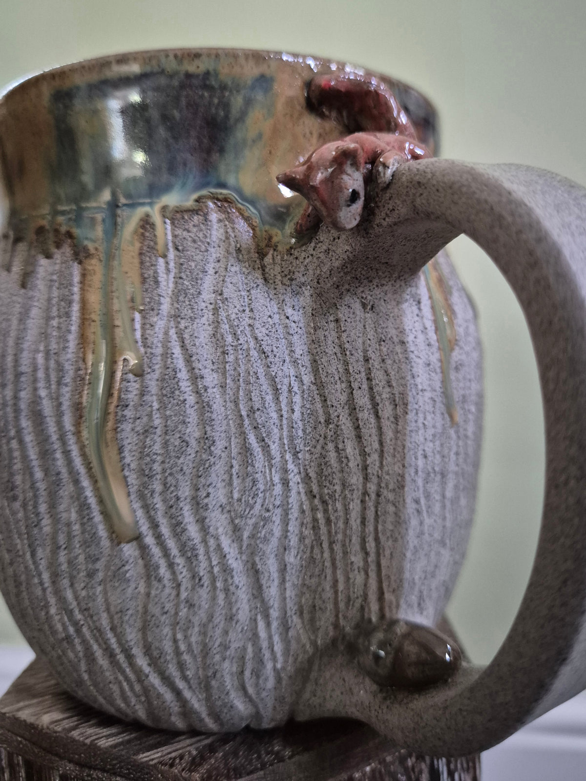 A close-up of a handmade ceramic mug with a textured, ribbed gray and beige body, a glossy blue-green glazed rim, and a hand-sculpted reddish-brown squirrel on top of the handlel, shown on a wooden surface.
