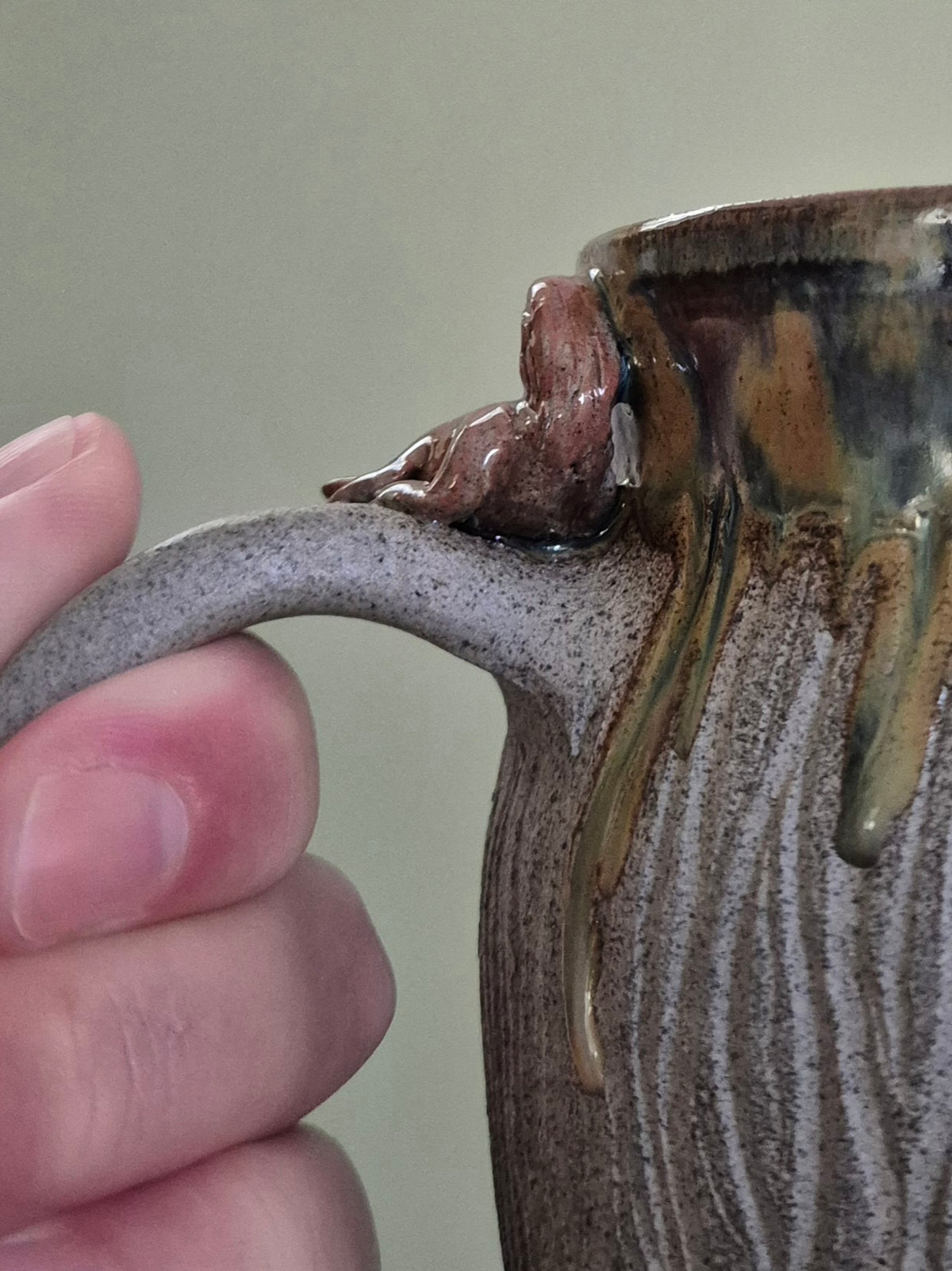 Close-up of a ceramic mug being held by a hand, showing a textured speckled gray handle and a glazed body with brown, gold, and green drip details.
