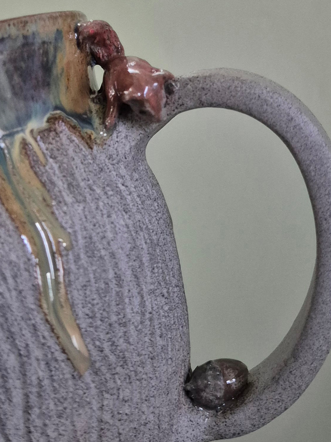 Close-up of a handmade ceramic pitcher with a textured speckled gray-purple body, a large handle, and small sculpted squirrel figure on the handle with an acorn at the base.