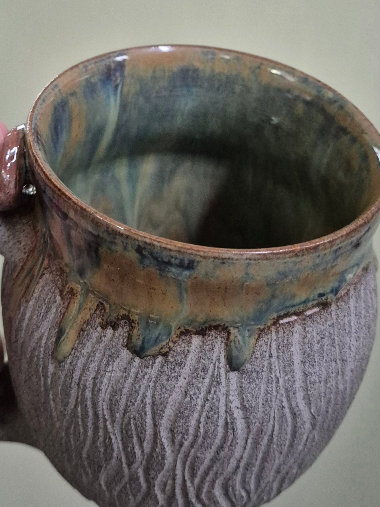 A close-up of a handmade ceramic mug with a textured, ribbed exterior in a speckled gray-brown glaze and dripping earthy brown and blue-green glaze along the rim.