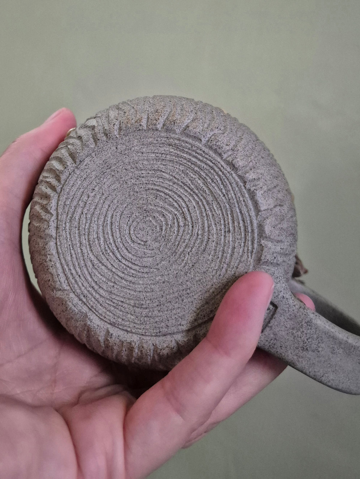 A hand holding a round, textured ceramic mug or coaster with a carved concentric circle pattern and a handle, shown against a muted olive-green background.