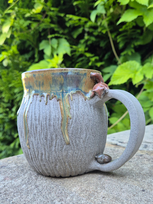 A handmade ceramic mug with a textured gray body, a rustic brown and gold dripping glaze rim, and a small sculpted squirrel on the handle, photographed outdoors on a stone surface with green foliage in the background.