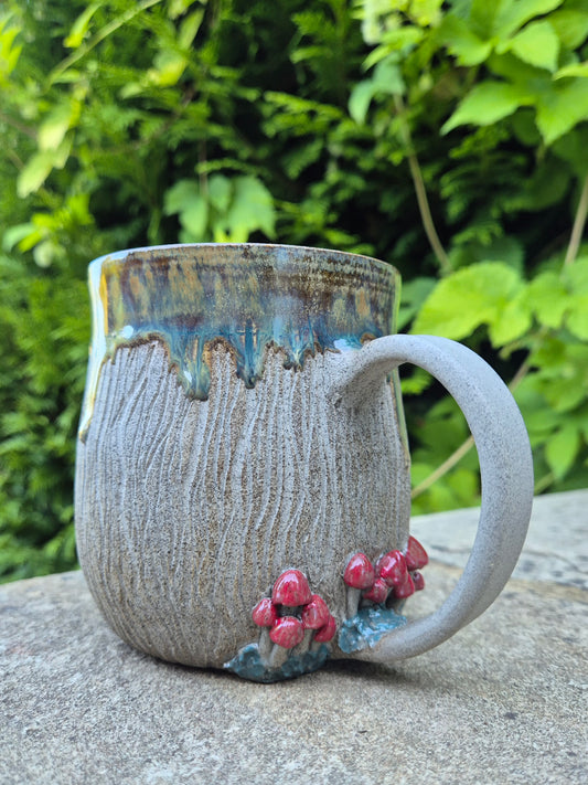 A handmade ceramic mug with a textured gray-brown body, a blue and gold drip glaze rim, and small red mushroom decorations near the base, sitting on a stone surface with a blurred green garden background.