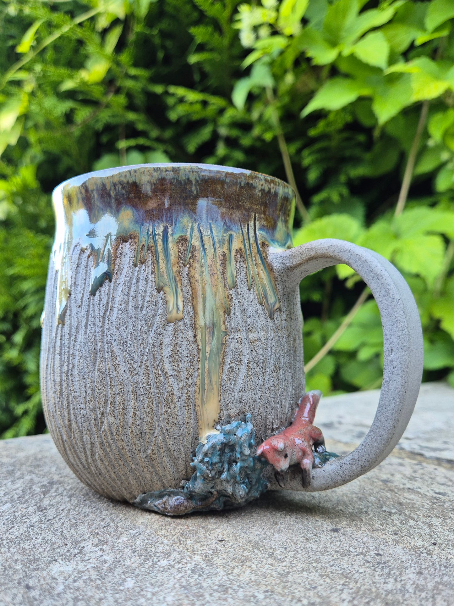 A handmade ceramic mug with a textured, ribbed gray-brown glaze, featuring a dripping multicolor design and a small 3D fox figure on the base, set outdoors on a stone surface with green foliage in the background.