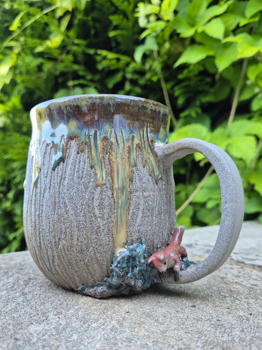 A handmade ceramic mug with a textured, ribbed gray-brown glaze, featuring a dripping multicolor design and a small 3D fox figure on the base, set outdoors on a stone surface with green foliage in the background.