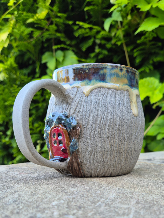 A handmade ceramic mug with a textured gray-brown glaze, featuring a small red fairy door on the side and a dripping gold and blue glazed rim, set outdoors on a stone surface with green foliage in the background.