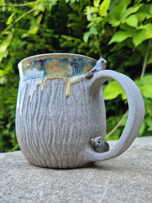 A handmade ceramic mug with a textured gray body, a blue and gold glazed rim, and small owls on the handle, shown outdoors on a stone surface with green foliage in the background.