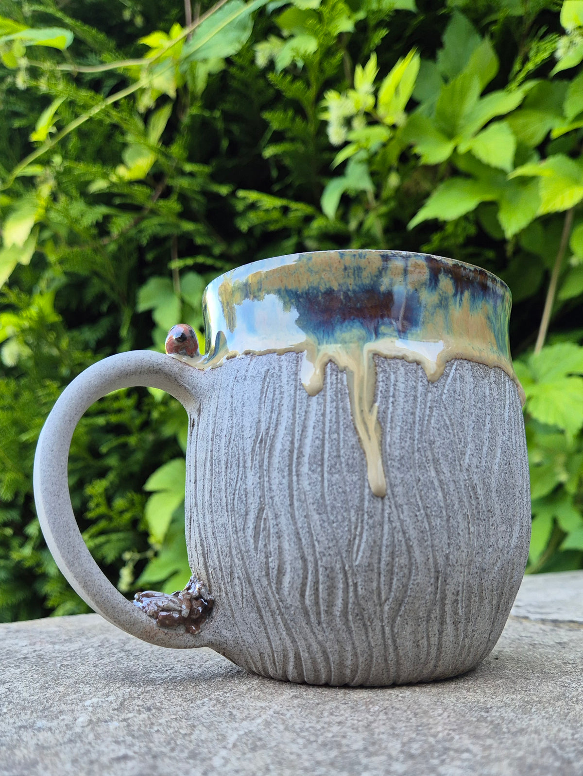 A handmade ceramic mug with a textured gray body, a dripping blue and gold glaze rim, and a small bird accent on the handle, set outdoors on a stone surface with green foliage in the background.
