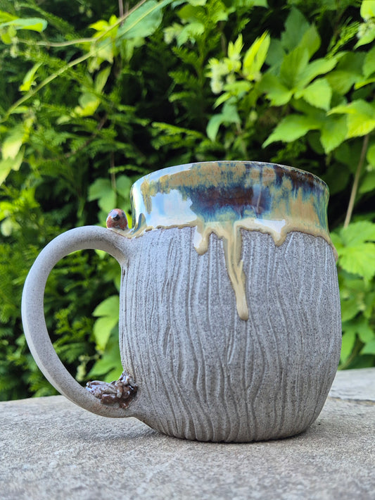 A handmade ceramic mug with a textured gray body, a dripping blue and gold glaze rim, and a small bird accent on the handle, set outdoors on a stone surface with green foliage in the background.