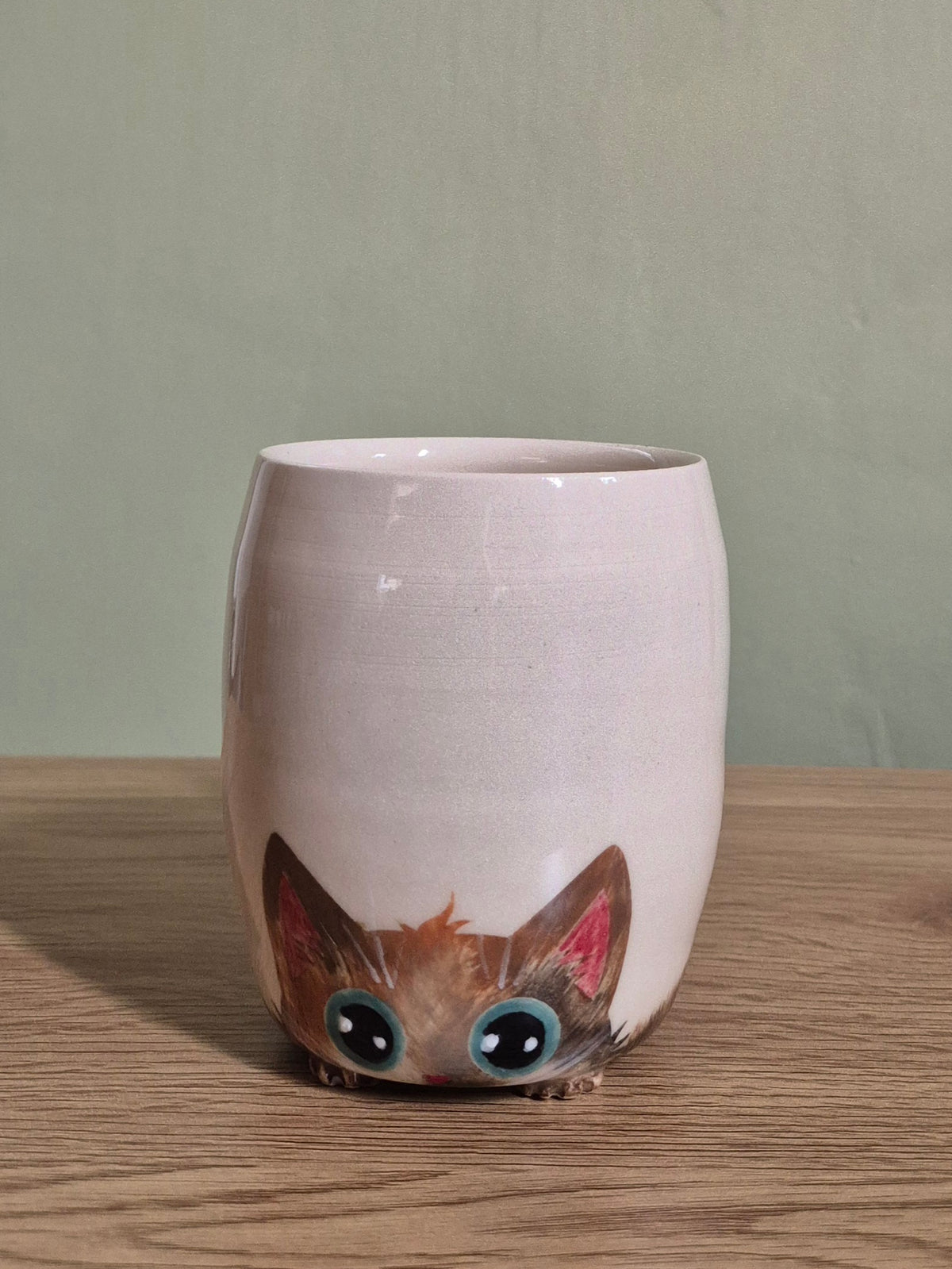 A light beige ceramic mug with a cute cartoon cat face peeking out from the bottom, sitting on a wooden surface against a muted green wall.