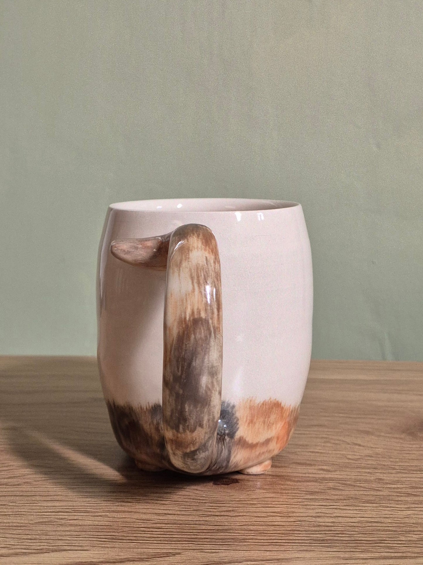 A ceramic cat mug with a glossy finish, featuring a sculptural tail handle with a natural brown fur pattern, sitting on a wooden table against a muted green wall.