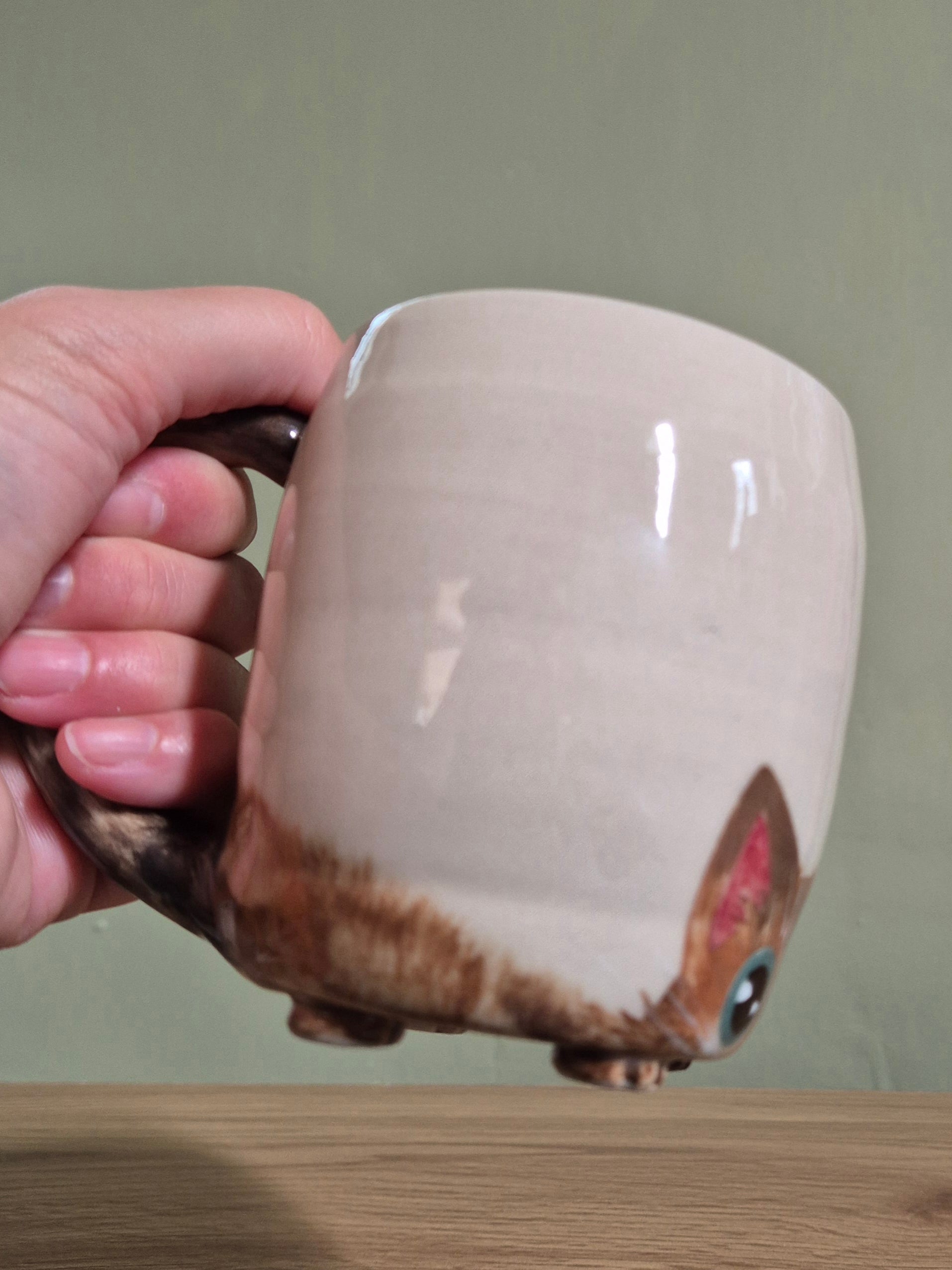 A hand holding a handmade ceramic mug with a cat design. The mug has a glossy beige body with a brown painted cat face and ears on the bottom right, and a dark brown handle.