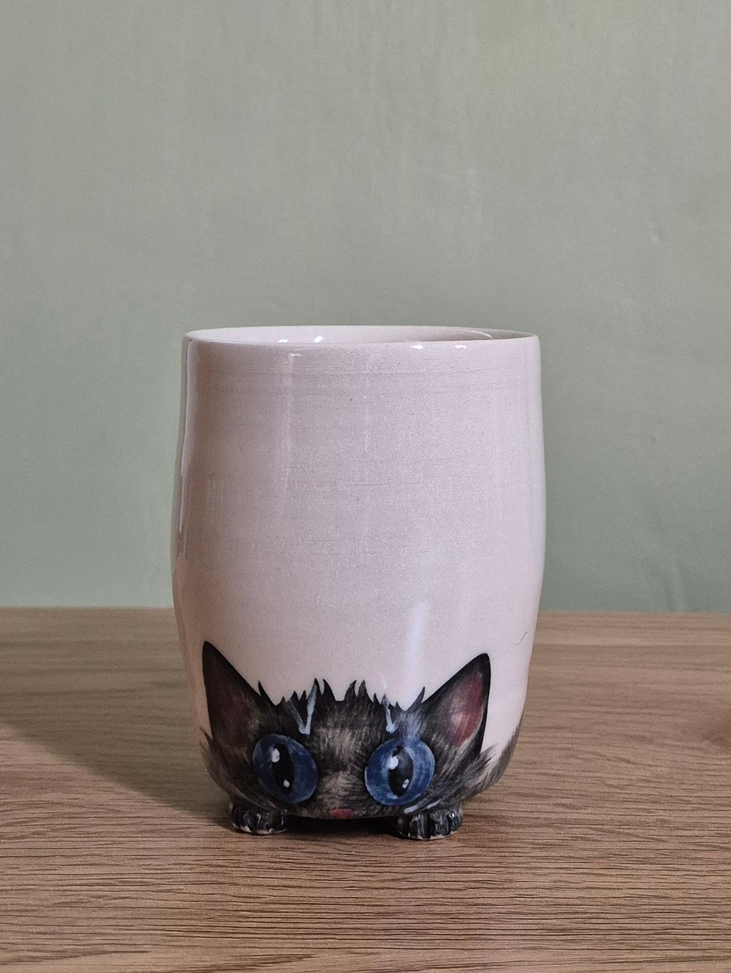 A light beige ceramic mug with a hand-painted black cat peeking out from the bottom, featuring large blue eyes, set on a wooden surface against a muted green wall.