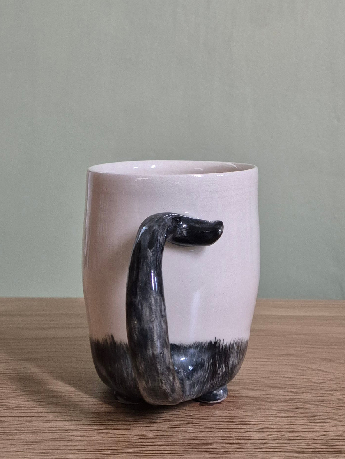 A handmade ceramic cat mug with a pale beige upper body, a glossy black textured base, and a sculptural tail handle , set on a wooden tabletop against a muted sage green background.