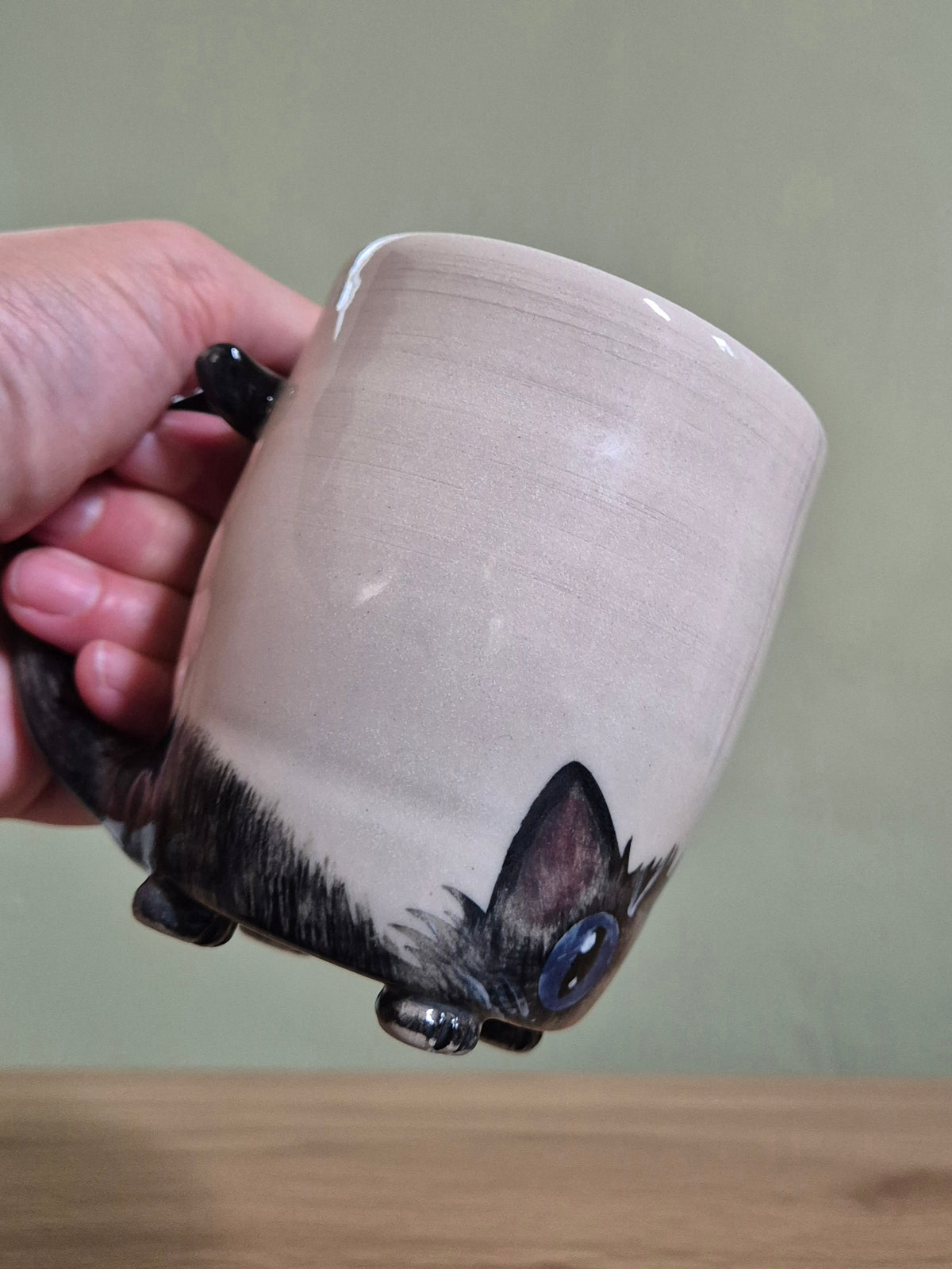 A hand holding a ceramic mug shaped like a cat peeking out from the bottom, with a beige upper body and a dark cat-face design on the base, against a muted green wall and wooden tabletop.