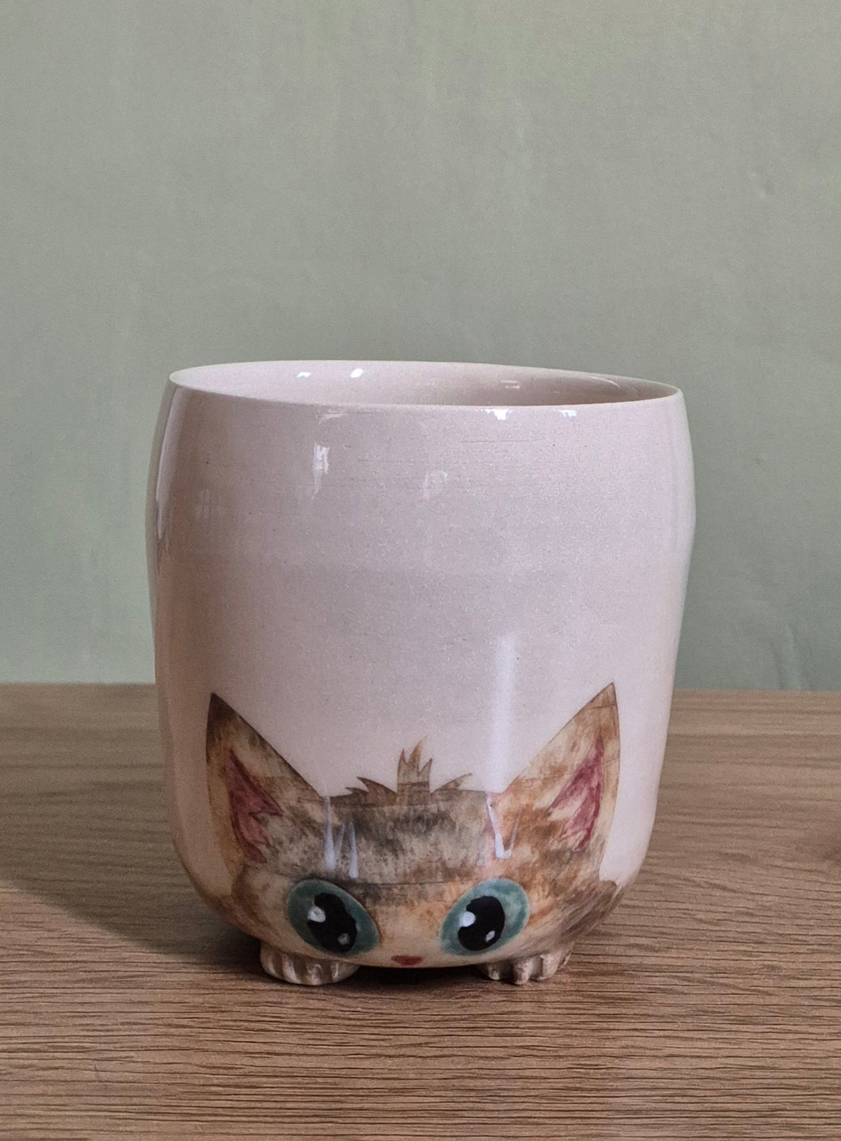 A light beige ceramic mug shaped like a cat's head, featuring large blue eyes and pointed ears, sitting on a wooden surface against a muted green wall.