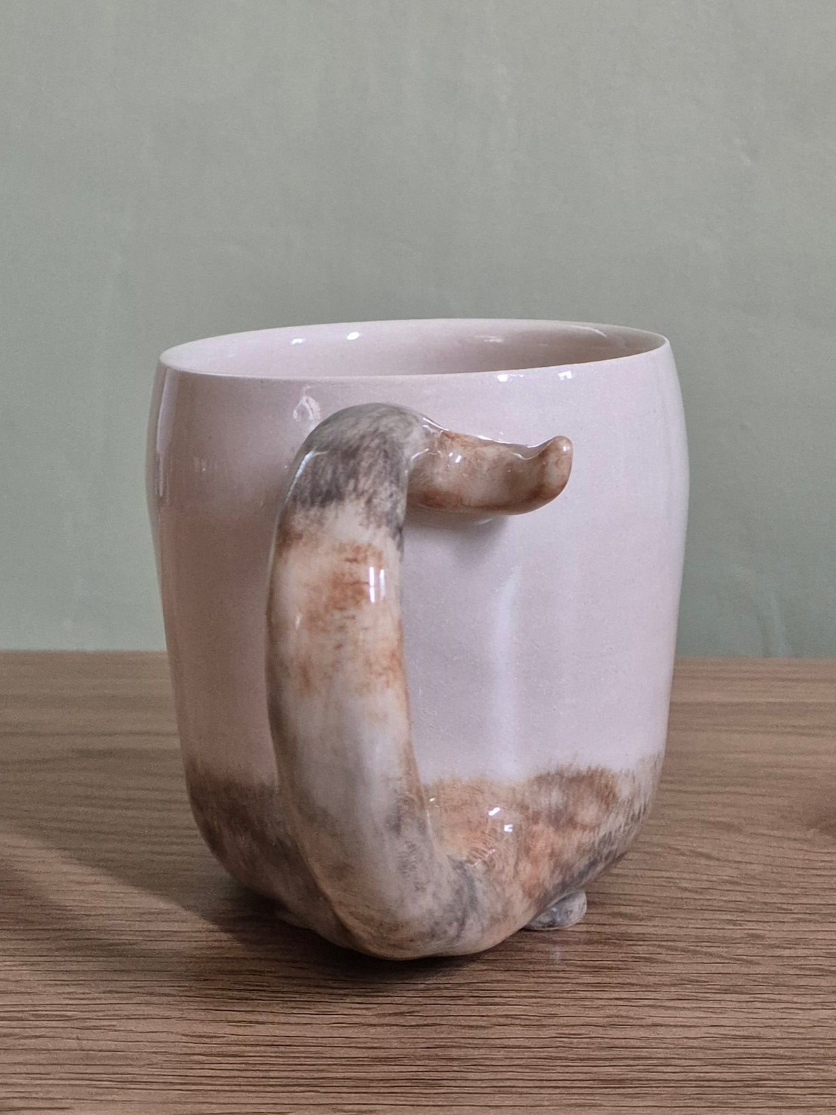 A beige ceramic mug with a unique sculptural handle resembling a cat's tail and hand-painted cat fur patterns. Set on a wooden tabletop against a muted green wall.