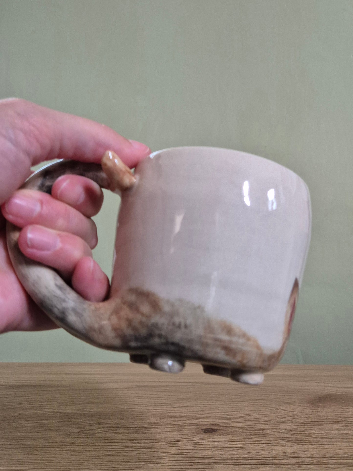 A handmade ceramic mug with a sculptural antler handle, held by a hand over a wooden tabletop against a muted green wall.