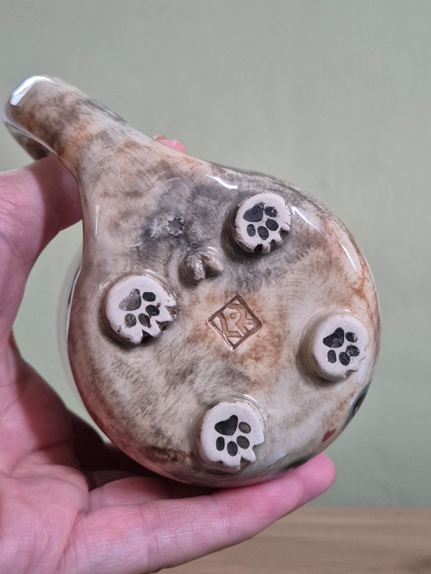 A hand holding a ceramic mug, showing the bottom of the mug with five raised paw-print motifs and a small stamped mark in the center, against a plain indoor background.