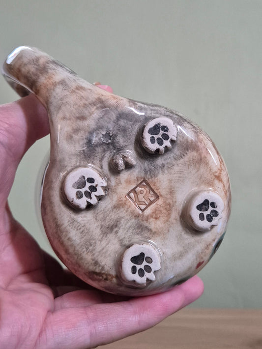 A hand holding a ceramic mug, showing the bottom of the mug with five raised paw-print motifs and a small stamped mark in the center, against a plain indoor background.