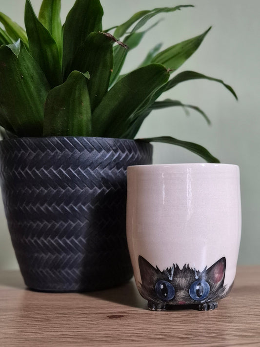 A light beige ceramic tumbler featuring a cute black cat with large blue eyes peeking out from the bottom, styled on a wooden table next to a potted green plant.