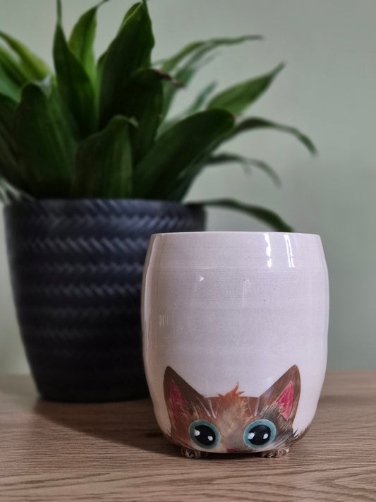 A light beige ceramic cup with a cute cartoon cat face peeking out from the bottom, styled on a wooden table with a blurred green potted plant in the background.