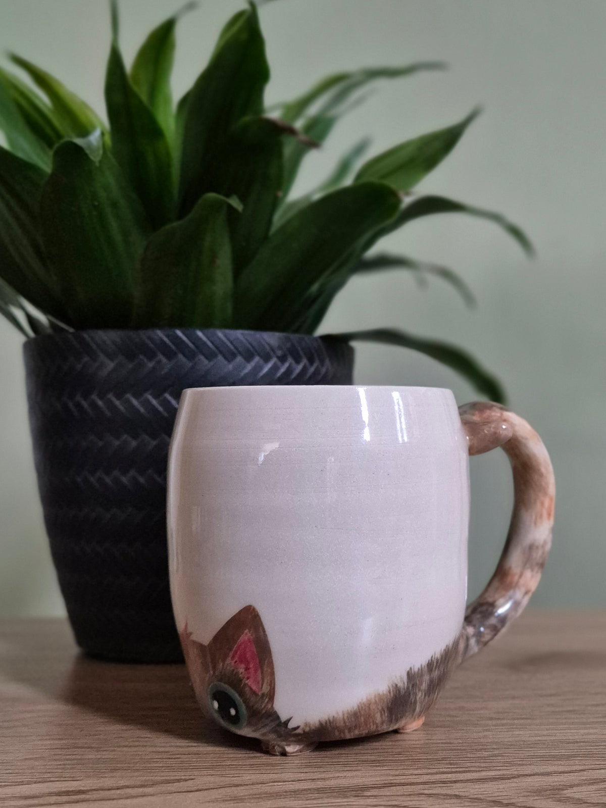 A ceramic mug with a pale beige body and a realistic cat body printed on the side, styled on a wooden table next to a potted green plant.