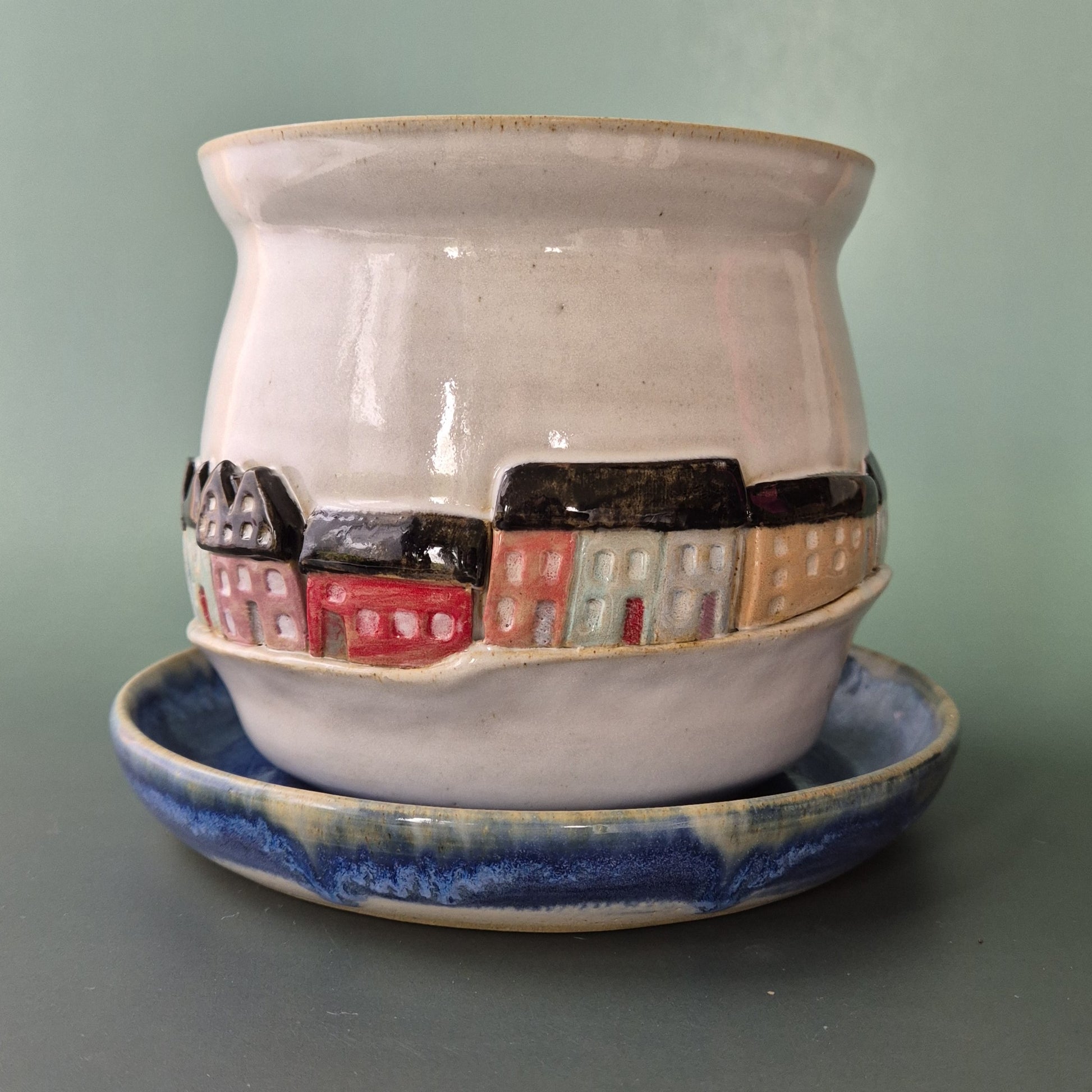 Ceramic pot with a cityscape design on a matching saucer against a green background