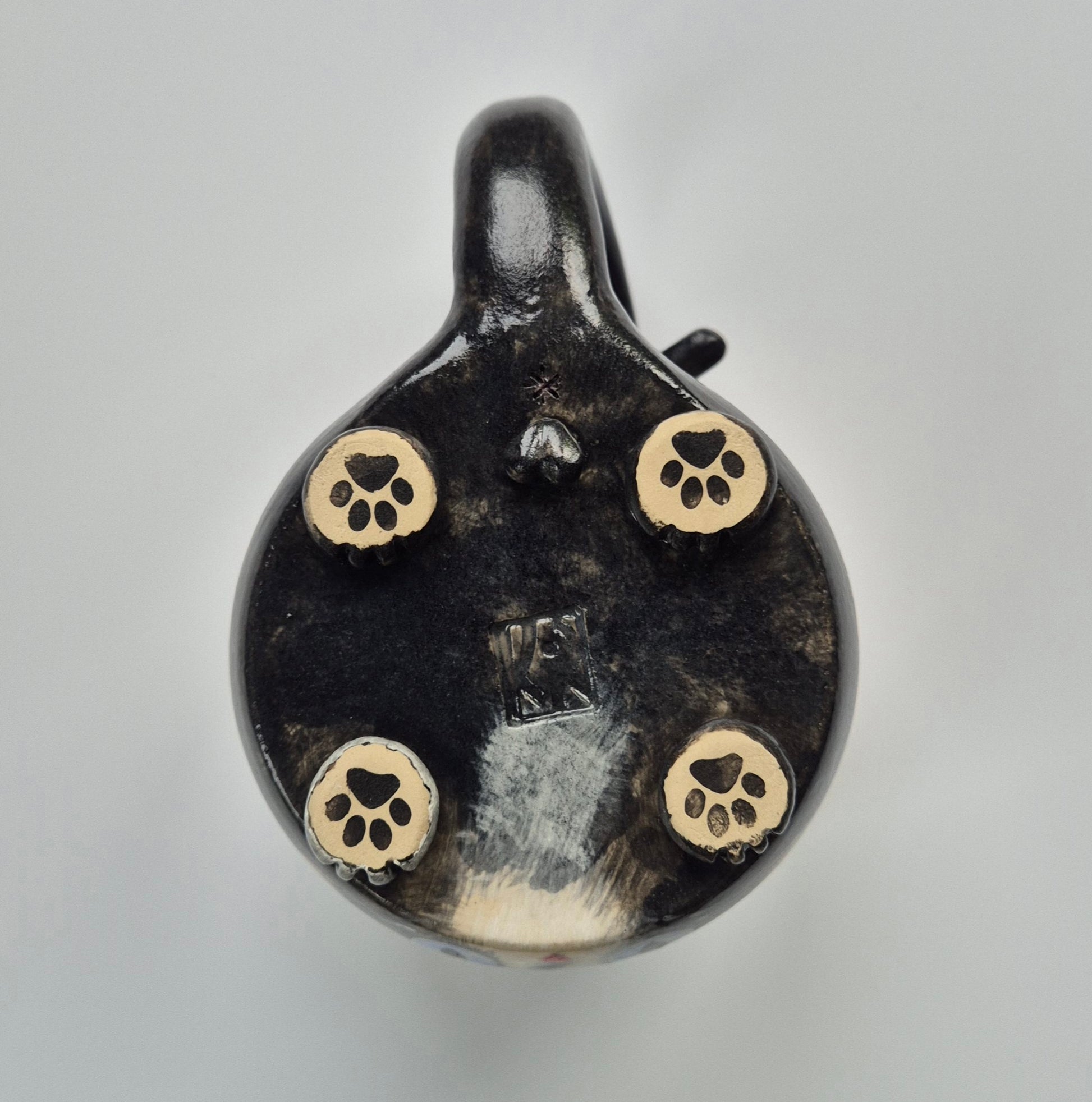 Base of a black ceramic cat mug with sculpted paws that have paw prints on a gray background. This mug also has balls (male cat)