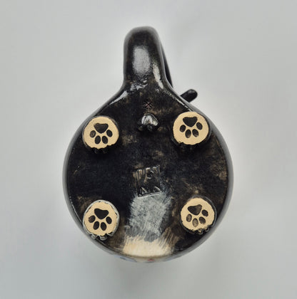 Base of a black ceramic cat mug with sculpted paws that have paw prints on a gray background. This mug also has balls (male cat)