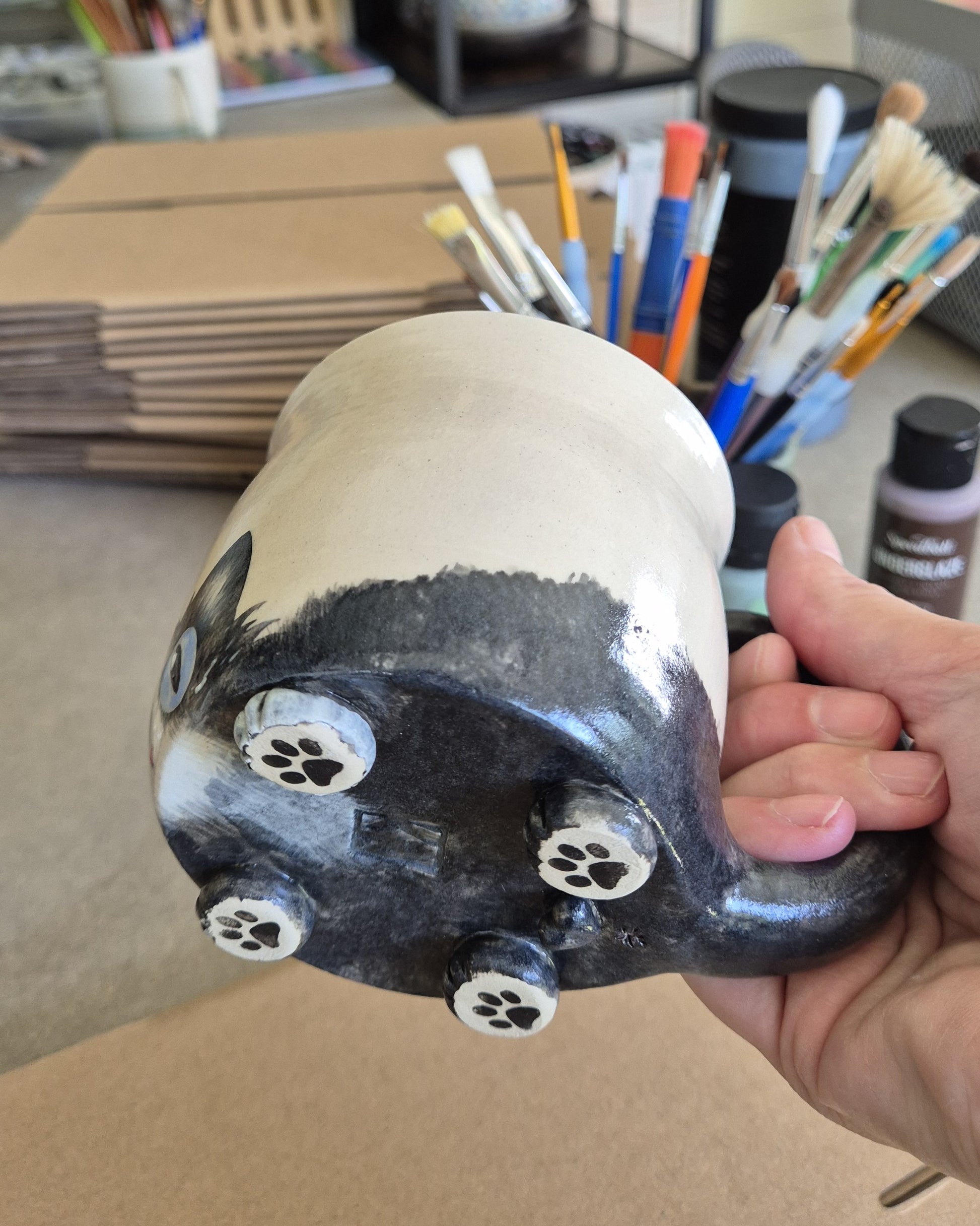 Hand holding a hand-painted ceramic cat mug, surrounded by art supplies.