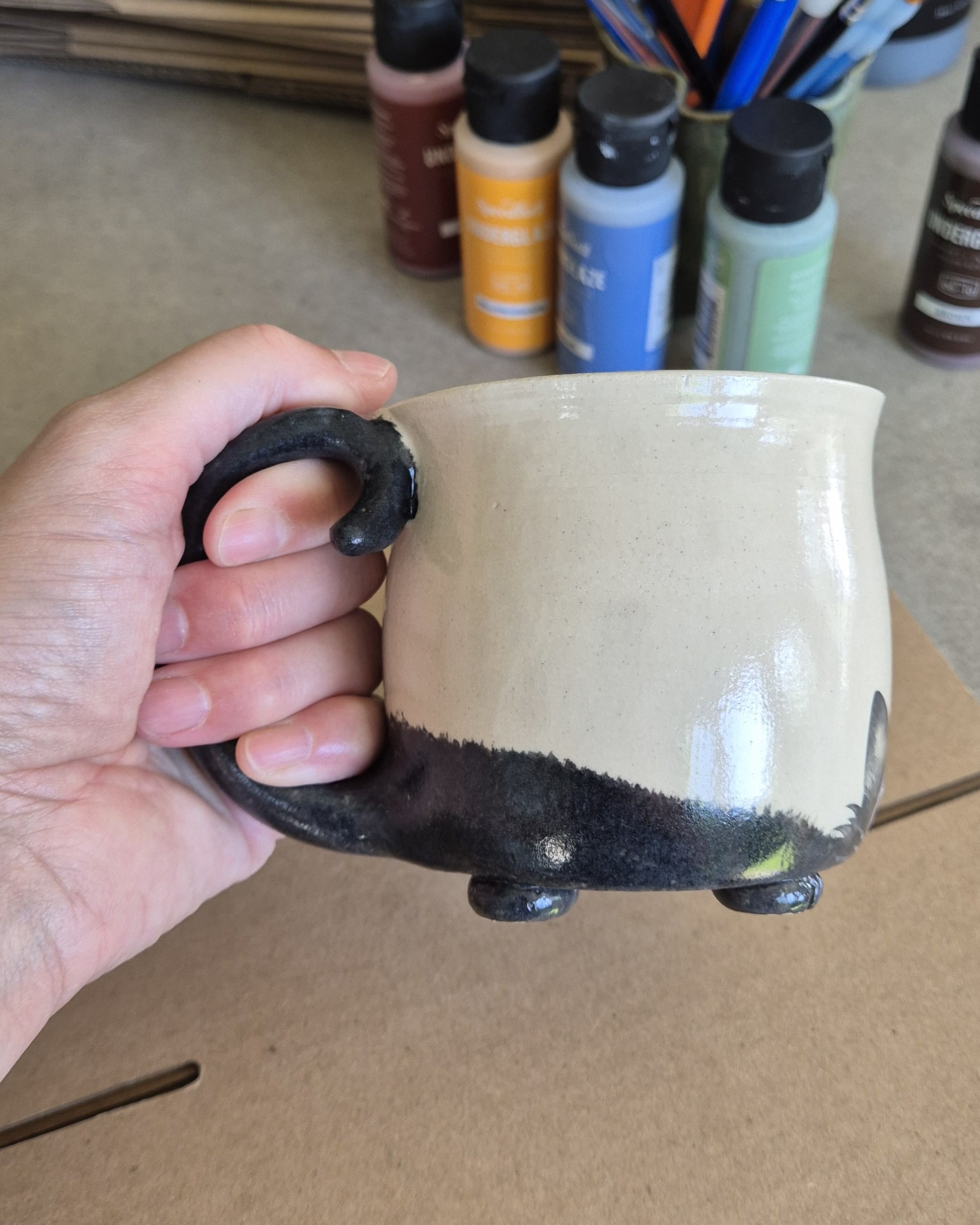 Hand holding a ceramic cat mug with black tail against a background of art supplies.