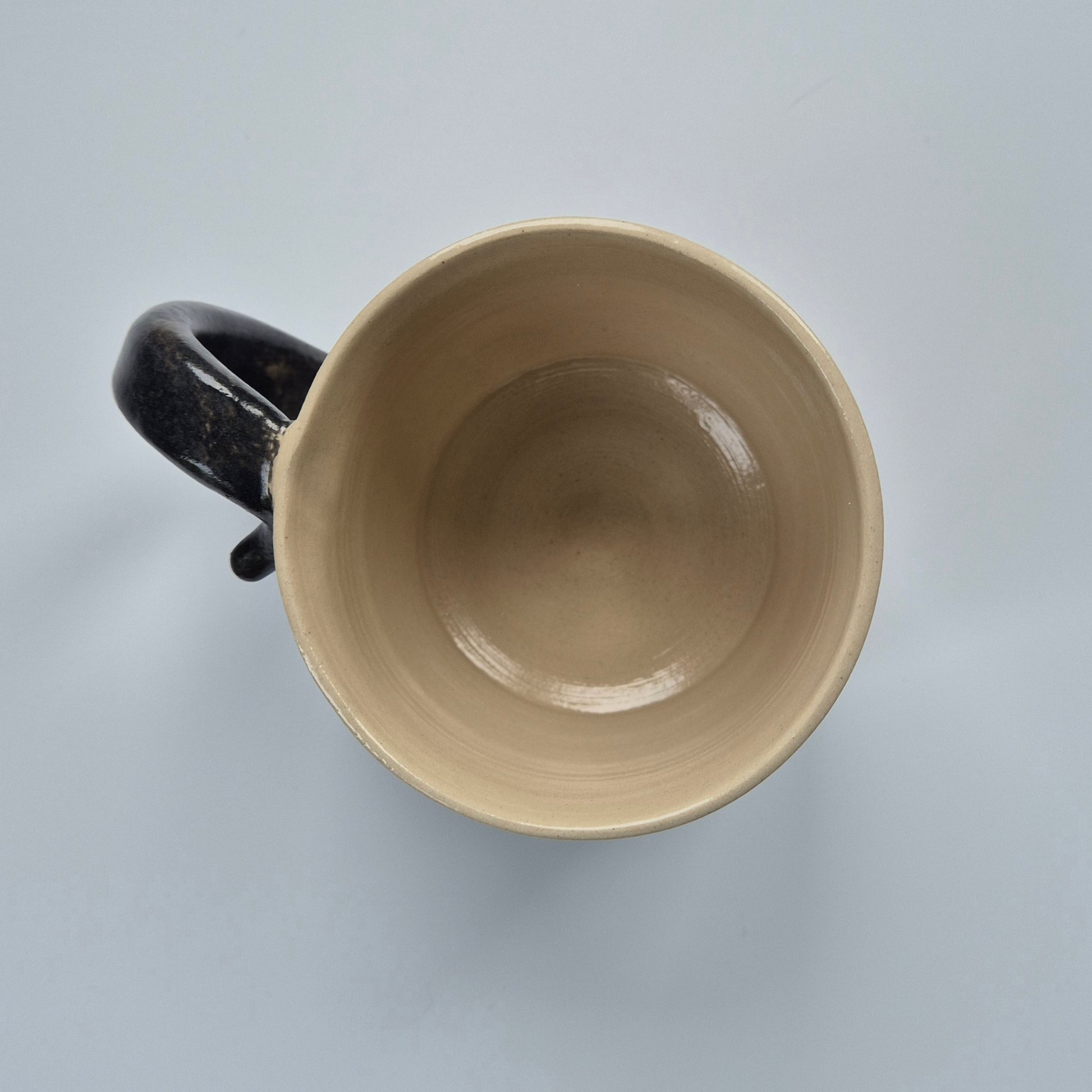 Top view of a ceramic cat mug showing the cream coloured inside. has a  black tail handle on a light gray background