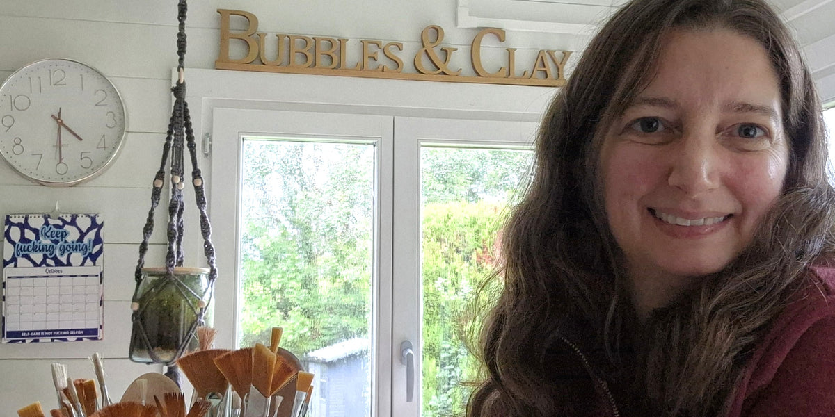 Kerry smiling in front of a window in her studio with 'Bubbles & Clay' sign and various items in the background.