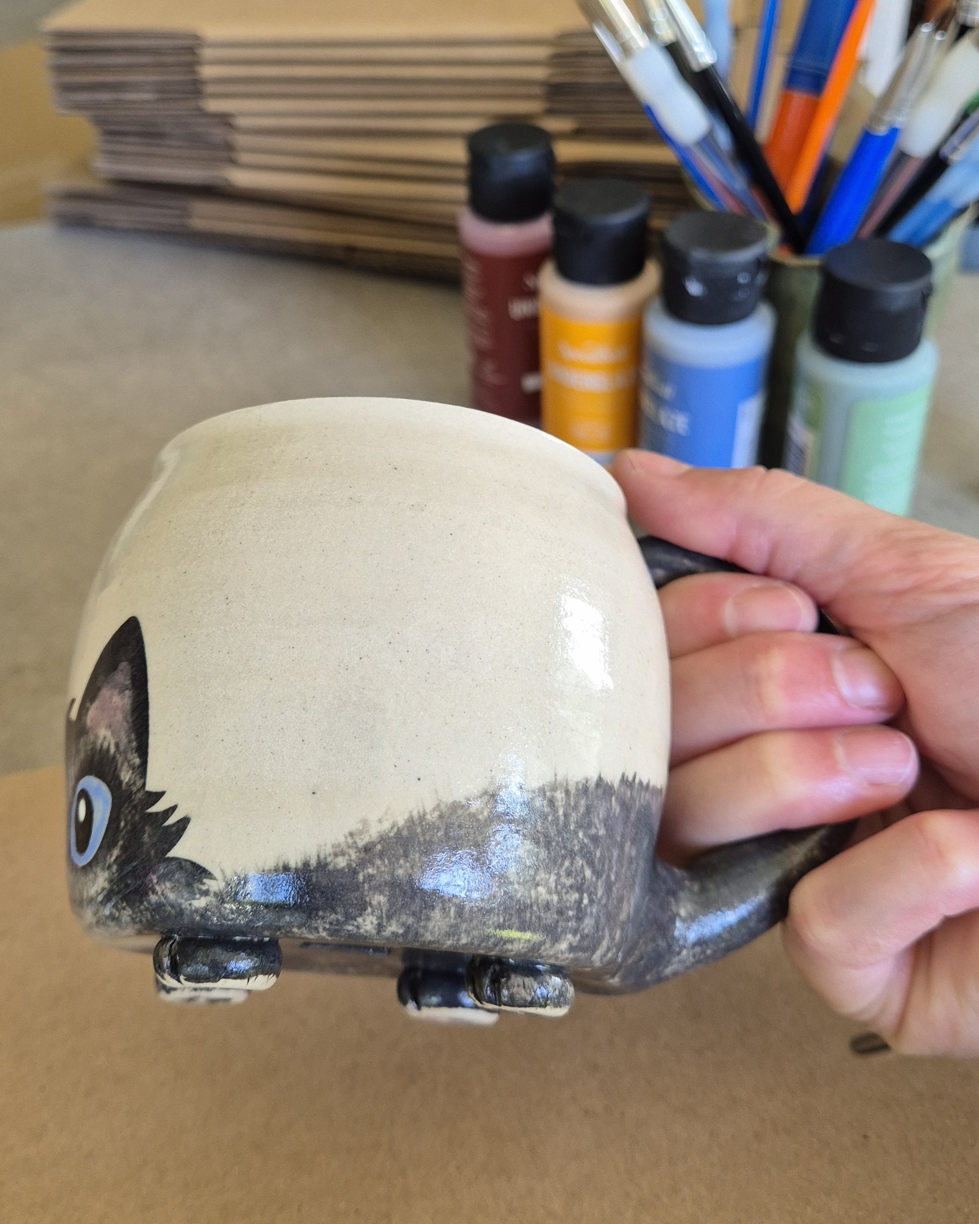 Hand holding a ceramic cat mug with paint supplies in the background