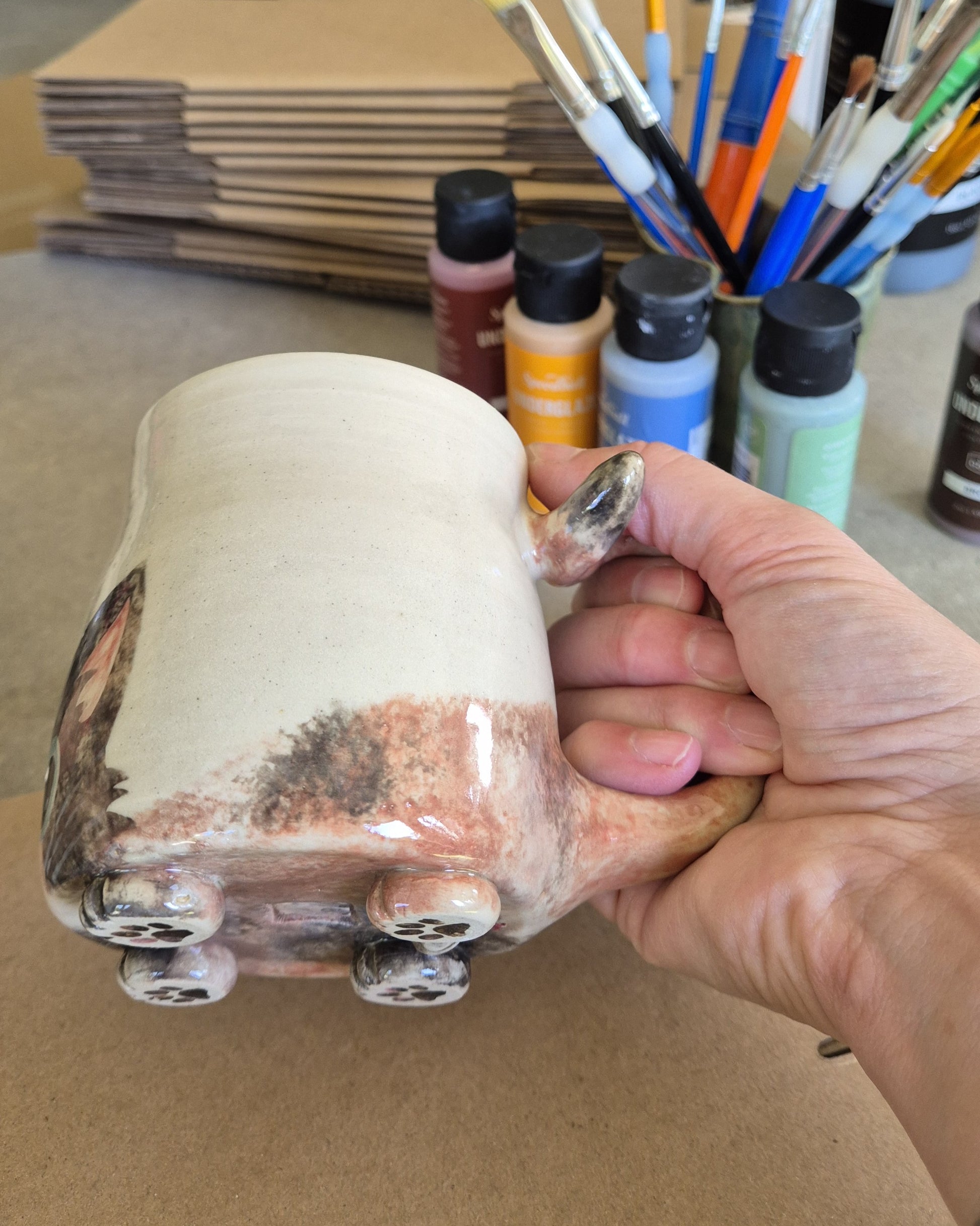 Hand holding a ceramic mug with painted features, surrounded by art supplies on a table.