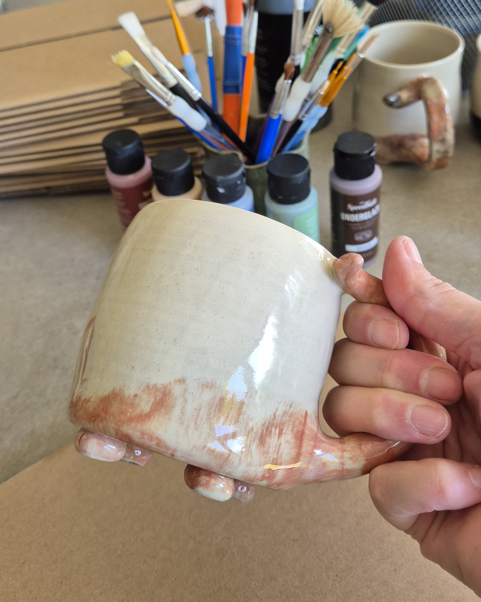 Hand holding a ceramic mug with paint supplies in the background