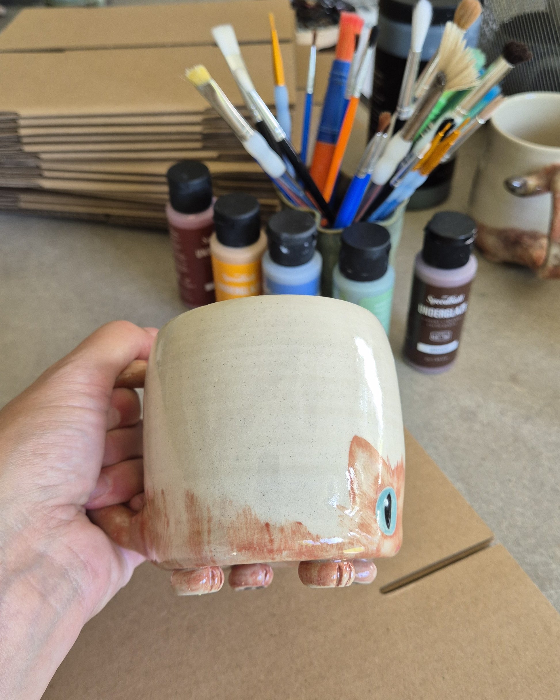 Hand holding a ceramic mug with paint bottles and brushes in the background