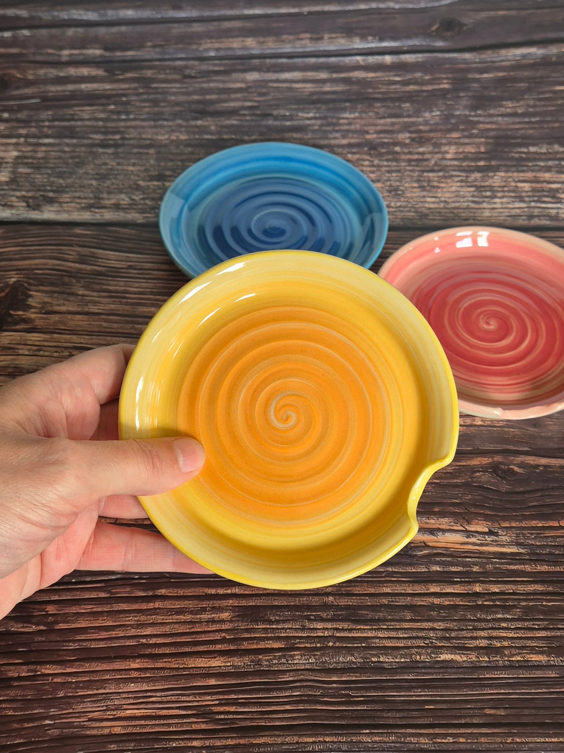 Yellow spoon rest held by a hand with blue and pink ones in the background on a wooden surface