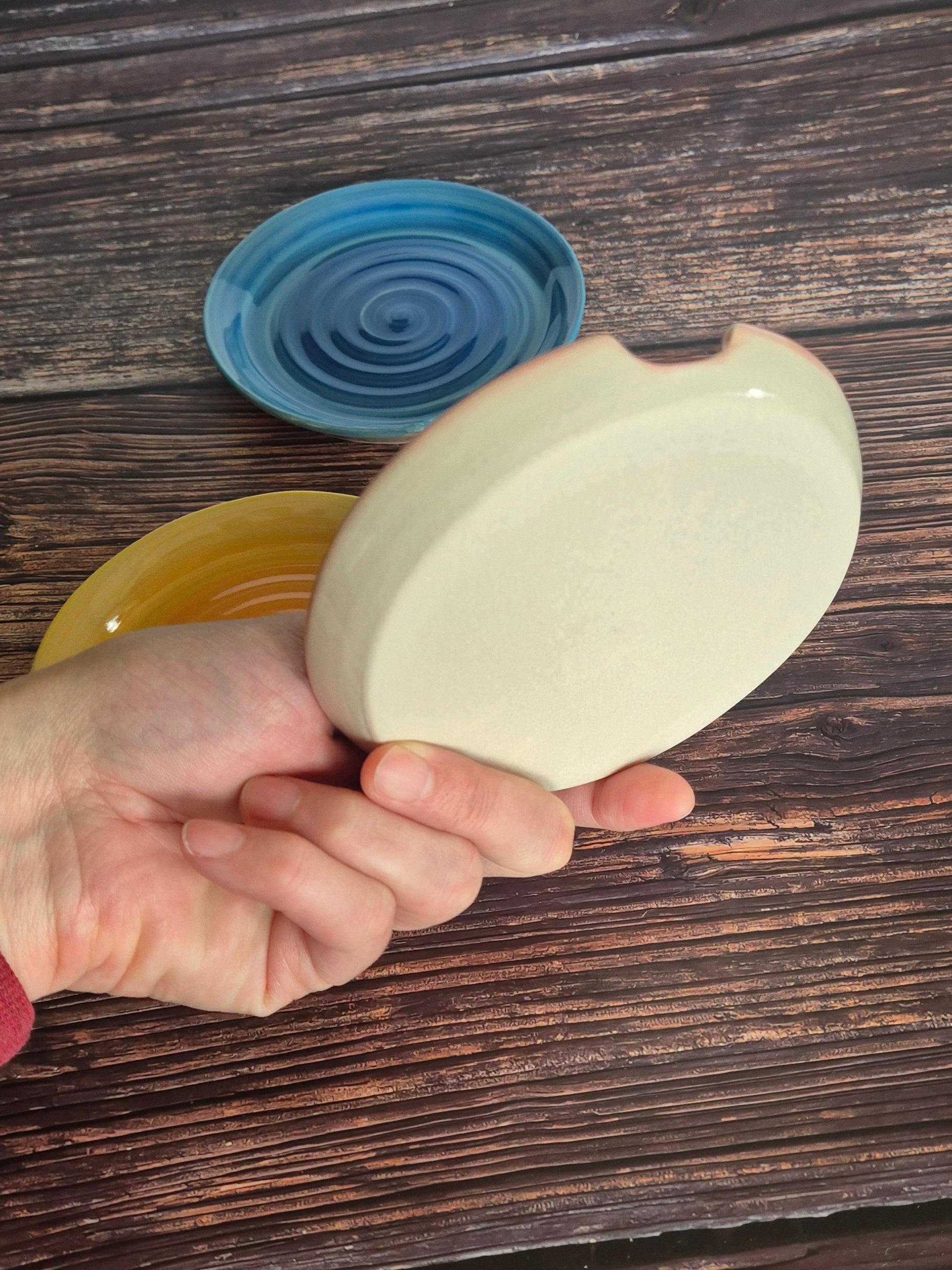 Hand holding a ceramic spoon rest to show the base with blue and yellow ones in the background on a wooden surface